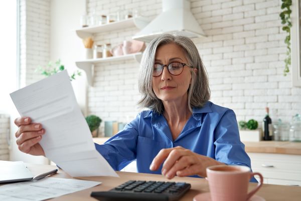 Woman sitting in kitchen with looking at a bill with a calculator and cup of coffee beside her.